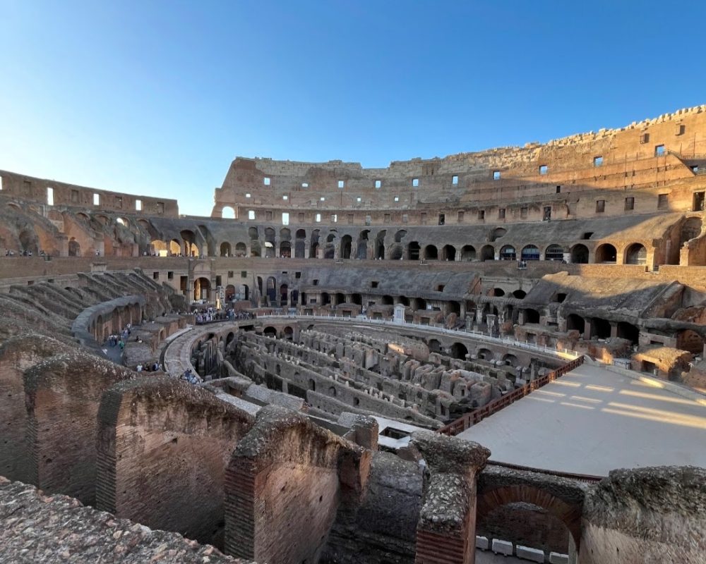 A panoramic view of the interior of the Colosseum in Rome, showcasing ancient stone architecture and rows of seats, illuminated softly by evening light.
