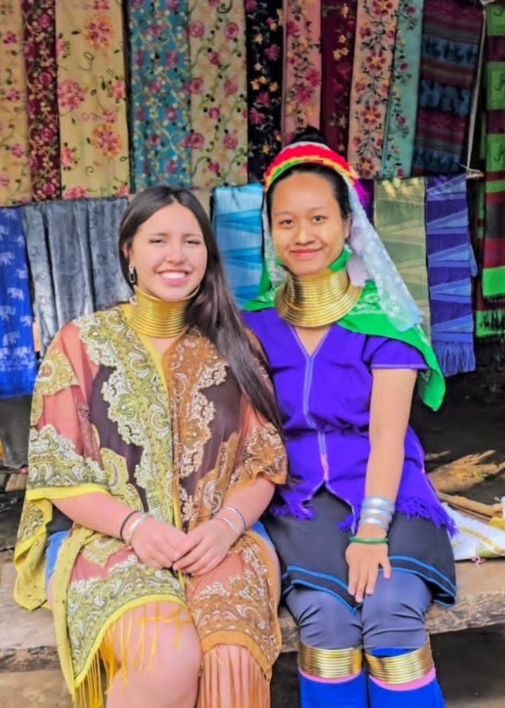 The White Temple, Chiang Rai Two women, one wearing a colorful shawl and the other in traditional Kayan attire with neck rings, sit together in front of vibrant textiles.