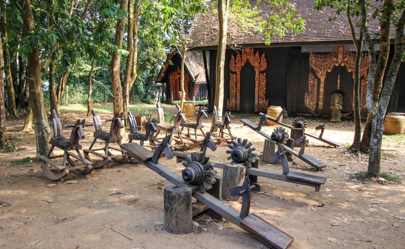 Baan Dam: The Black House Museum A collection of handcrafted wooden rocking horses and balance toys arranged in a shaded area surrounded by trees, with traditional architecture visible in the background.